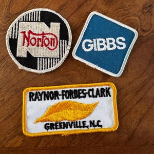 Puede incluir: Tres parches bordados con logotipos de empresas. Uno es un círculo blanco y negro con "Norton" en rojo. Otro es un rombo azul con "GIBBS" en blanco. El tercero es un rectángulo blanco con "RAYNOR-FORBES-CLARK GREENVILLE, N.C." y una hoja dorada.
