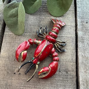 May include: A red lobster brooch with gold accents and black claws. The brooch has two small green gems and is set against a rustic wooden background. The lobster's body is painted red with gold trim.