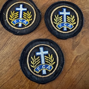 Puede incluir: Tres parches circulares con fondo negro. Cada parche presenta una cruz blanca, una pancarta azul con el texto "FCA" y un diseño dorado en forma de trigo. Los parches son probablemente para ropa o accesorios.