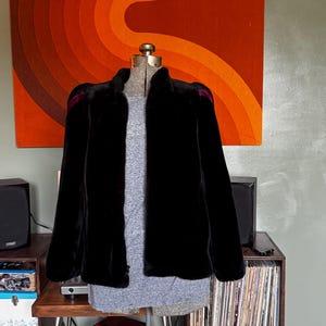 May include: A black faux fur jacket with a high collar and long sleeves, featuring dark purple velvet accents on the shoulders. The jacket is displayed on a dress form over a grey heathered t-shirt, showcasing a structured fit and soft texture.
