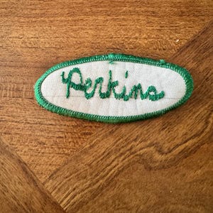 May include: An oval-shaped embroidered patch featuring a white fabric background with a green stitched border. The word Perkins is embroidered in green cursive script across the center of the patch, which has a clean, finished edge design.