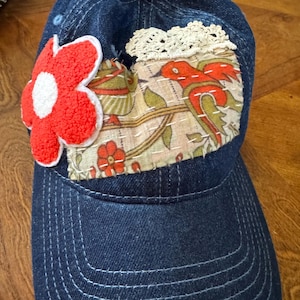 May include: A dark blue denim baseball cap featuring a large, textured red and white flower appliqué on the left side. A rectangular fabric patch with a bird and floral print is stitched to the front, accented by a white crocheted lace trim.