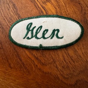 May include: An oval-shaped embroidered patch featuring a white fabric background with a dark green stitched border. The name Glen is embroidered in the center in a dark green cursive script font, creating a clear contrast against the white base.