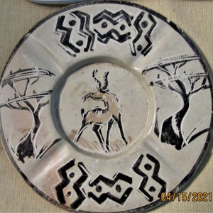 May include: A white ceramic plate with a black and white design. The design features a stylised antelope, two trees, and a geometric pattern. The plate has a raised rim and a shallow well in the centre.