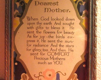 May include: A framed vintage print with the text "Dearest Mother." The print features an ornate design with floral and leaf motifs in shades of brown, blue, and yellow. The frame is a dark brown with a blue accent.