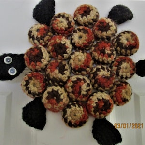 Puede incluir: Un posavasos de tortuga tejido a crochet con una concha marrón, naranja y beige. La tortuga tiene patas de fieltro negro y una cabeza de fieltro negro con dos ojos saltones blancos.