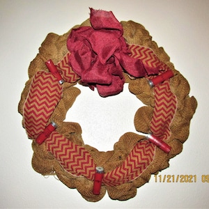 May include: A brown burlap wreath decorated with red and gold chevron ribbon and red shotgun shells. The wreath has a large red bow in the center.