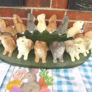 May include: A collection of ceramic bunny figurines in various colors, including tan, gray, and cream, arranged on a green, leaf-shaped base. The Easter-themed display is set against a brick background and a blue and white checkered tablecloth.