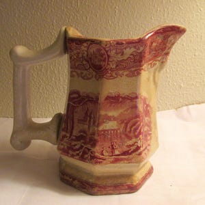 May include: A vintage pitcher with a cream-colored base and a red landscape design. The pitcher has a unique octagonal shape and a curved handle. The red design features a scenic landscape with figures and architectural elements.