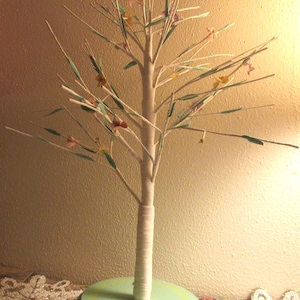 May include: A decorative tree sculpture with a light green base and a white trunk wrapped in twine. The branches are adorned with small, colorful paper leaves and flowers, creating a whimsical, nature-inspired accent piece.