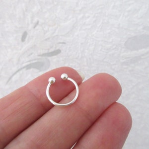 Peut inclure: Un petit anneau de nez argenté en forme de fer à cheval avec deux petites boules aux extrémités. Le bijou est tenu entre les doigts sur un fond blanc avec un motif subtil.