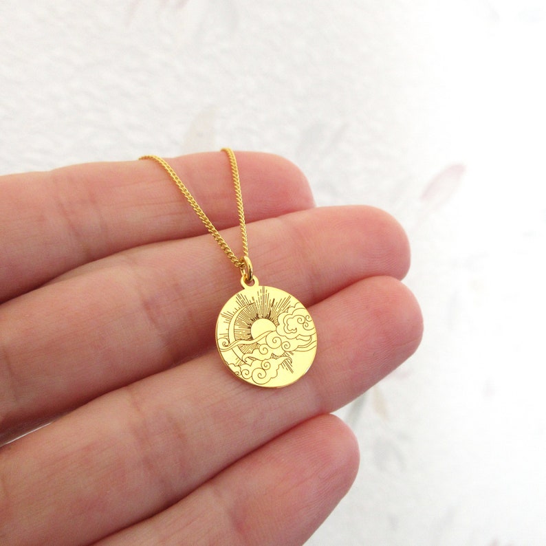 May include: Gold necklace with a round pendant featuring an engraved design of a sun and clouds.