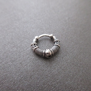 Può includere: Un piccolo orecchino a cerchio color argento con dettagli intricati. L'orecchino presenta una trama intrecciata e piccoli abbellimenti rotondi. Il design ha un'estetica antica o tribale, adatta a vari stili.