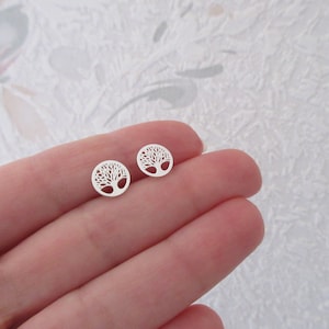 Puede incluir: Dos pendientes de botón de color plateado. Cada pendiente presenta un diseño circular con una silueta detallada de árbol de la vida. Los pendientes son pequeños y delicados, perfectos para el uso diario. Los pendientes se muestran en los dedos de una persona.