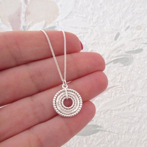 Peut inclure: Un collier en argent avec un pendentif circulaire composé de plusieurs anneaux concentriques. Le pendentif a un design texturé et perlé.