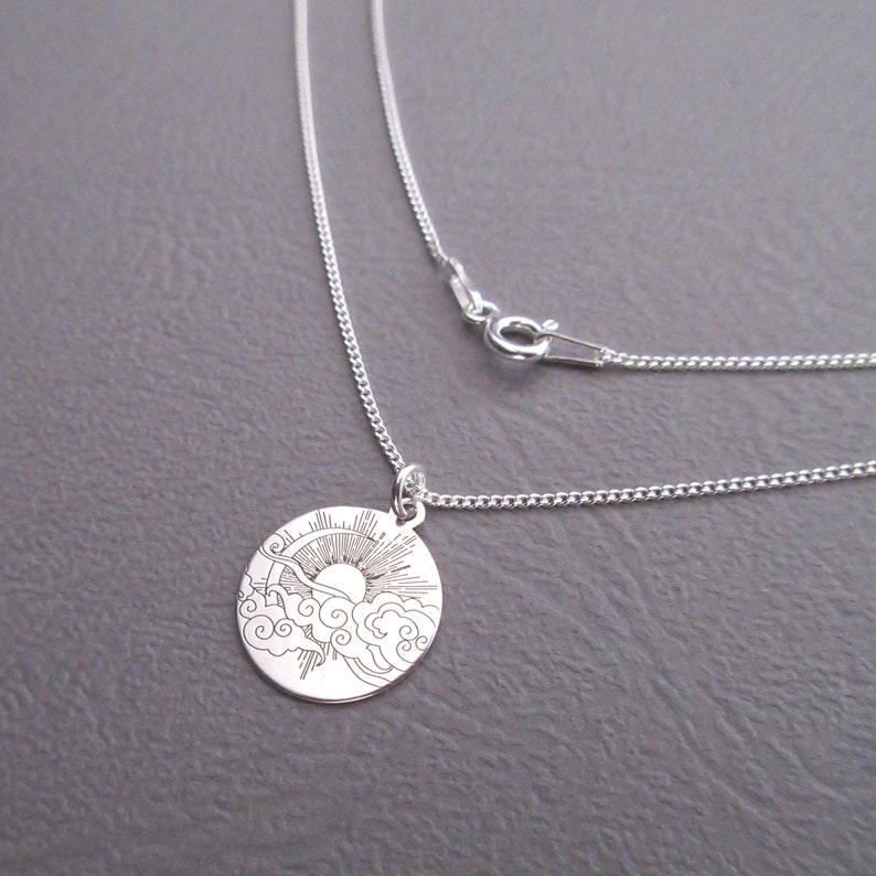 May include: A silver necklace with a round pendant featuring an engraved design of a sun rising above clouds.