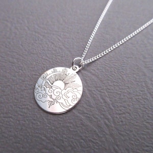 May include: A silver necklace with a round pendant featuring a sun and clouds design. The pendant is engraved with a detailed image of a sun rising above clouds.