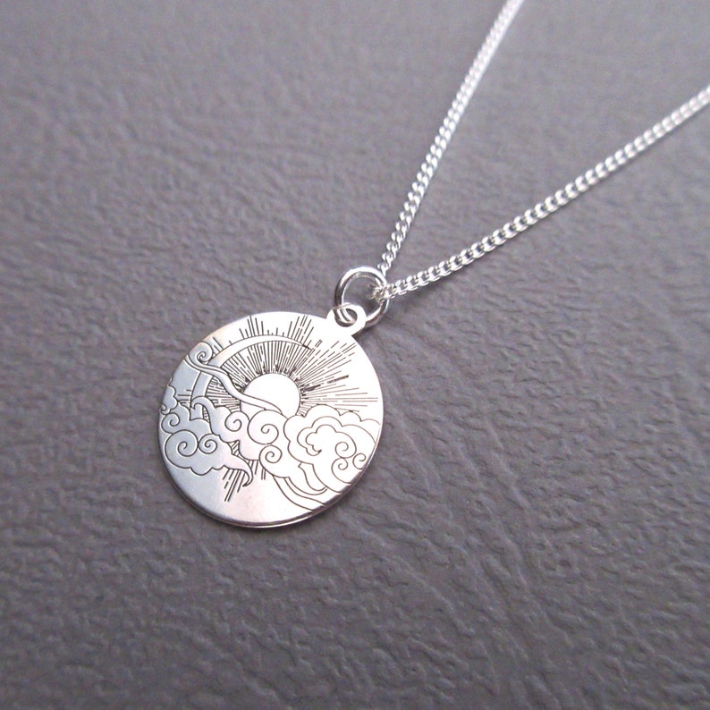 May include: A silver necklace with a round pendant featuring a sun and clouds design. The pendant is engraved with a detailed image of a sun rising above clouds.