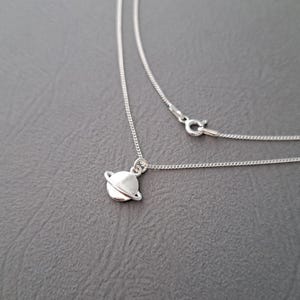 Peut inclure: Un collier en argent avec un petit pendentif de planète détaillé. Le collier est sur un fond gris.