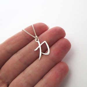 Peut inclure: Un collier avec un pendentif argenté en forme de lettre "P" stylisée. Le pendentif est suspendu à une fine chaîne. Le collier est présenté sur un fond clair.