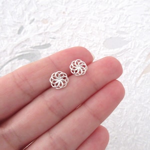 Op de afbeelding: Een paar kleine, zilverkleurige oorstekers. Elke oorbel heeft een delicaat, open ontwerp dat lijkt op een bloem of een ster. De oorbellen zijn rond en hebben een subtiele, reflecterende glans, wat wijst op een gepolijste metalen afwerking.