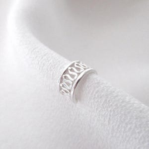 Peut inclure: Un ear cuff argenté avec un motif ajouré décoratif. Le bijou, une bande ornée d'un motif répétitif de lignes courbes, s'enroule autour de l'oreille. Le bijou est présenté sur un fond blanc.