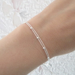 Könnte beinhalten: Ein zartes Silberkettenarmband mit kleinen Perlen. Das Armband besteht aus zwei übereinanderliegenden Ketten, was einen schlichten und eleganten Look erzeugt.