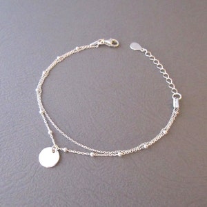 Op de afbeelding: Een zilveren kettingarmband met een kleine ronde hanger. De armband heeft twee kettingen, waarvan één ketting kleine kralen heeft.