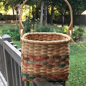 May include: A woven basket with a natural wood handle and a colorful woven pattern in shades of green, brown, and orange. The basket is hanging from a wooden fence.