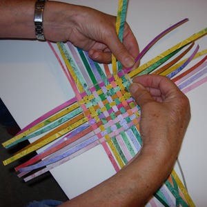 May include: A person weaving colourful paper strips together to create a basket. The strips are woven in a chequered pattern, creating a colourful and intricate design.