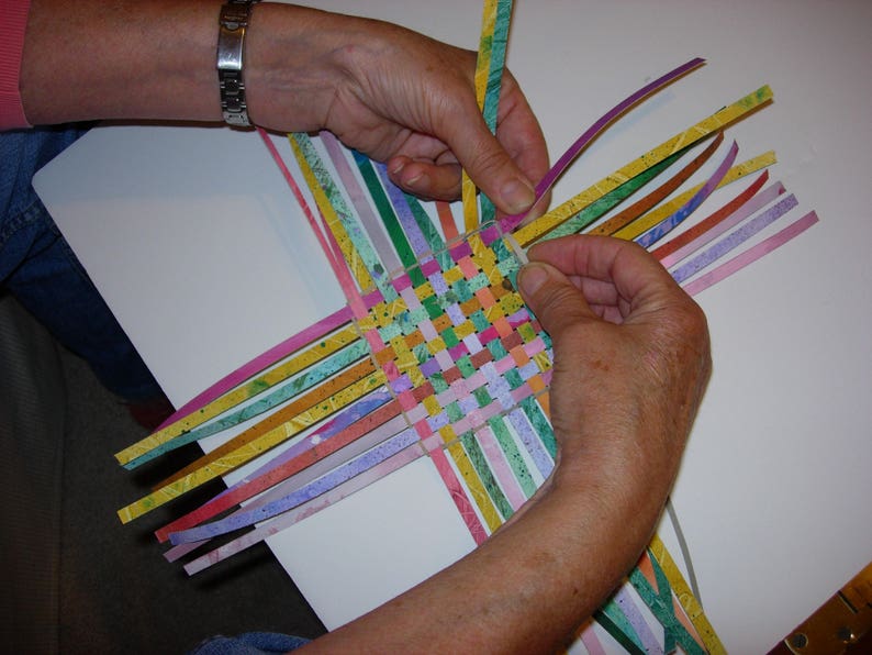 May include: A person weaving colourful paper strips together to create a basket. The strips are woven in a chequered pattern, creating a colourful and intricate design.