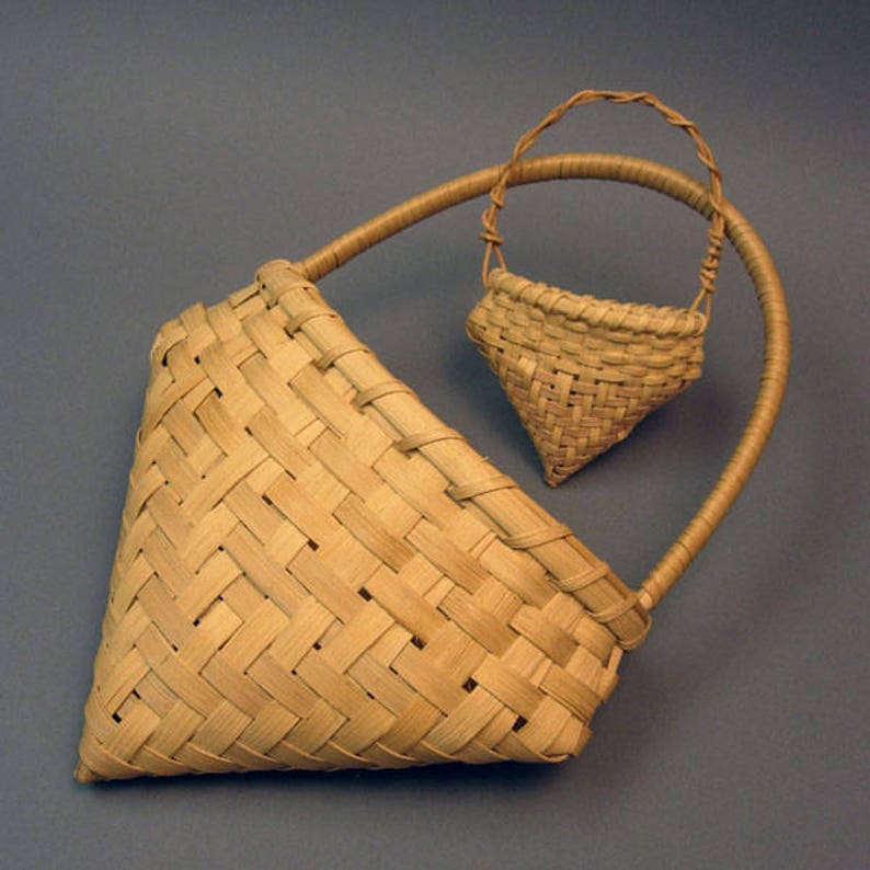 May include: Two woven baskets made of natural materials. The larger basket is triangular in shape with a handle. The smaller basket is hanging from the handle of the larger basket.