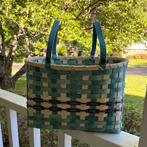 May include: A woven basket with blue and white stripes and a black trim. The basket has two blue handles with buttons. The basket is sitting on a white railing.