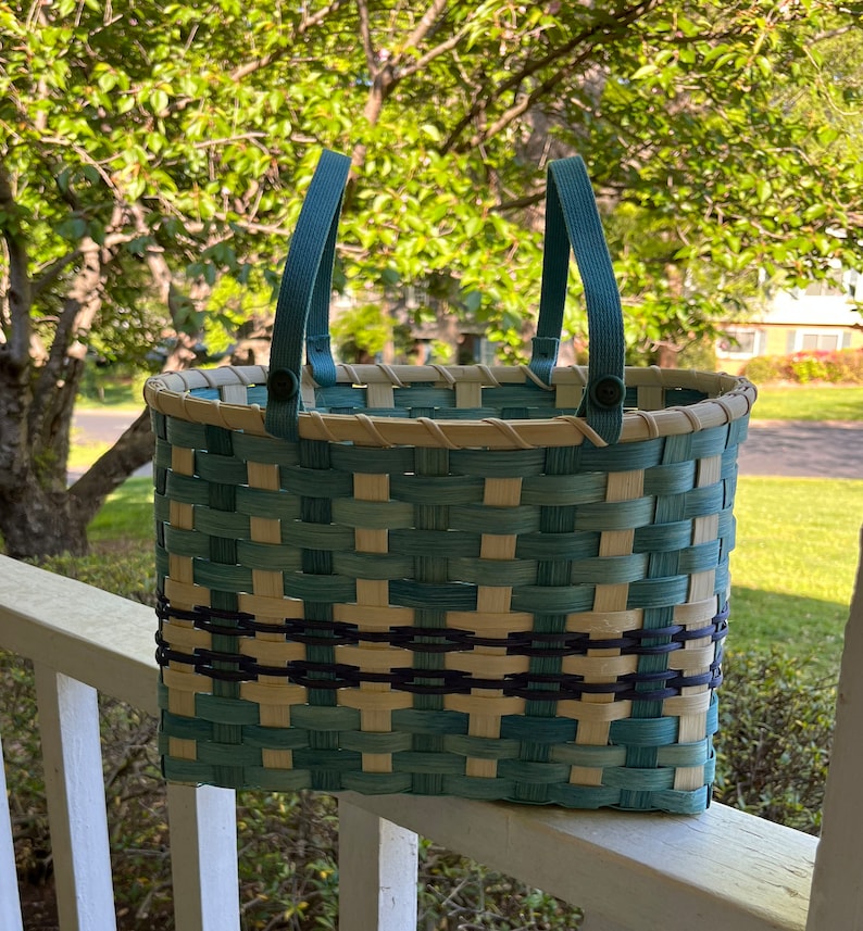 May include: A woven basket with blue and white stripes and a black trim. The basket has two blue handles with buttons. The basket is sitting on a white railing.