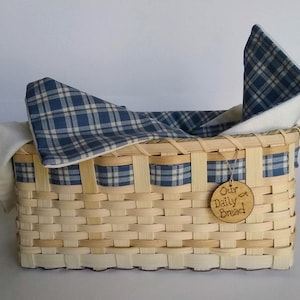 Puede incluir: Una cesta tejida con un forro de cuadros azules y blancos. La cesta tiene una etiqueta de madera que dice "Nuestro Pan Diario" con un pequeño símbolo de pez.