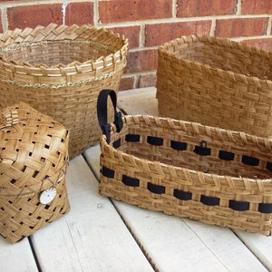 May include: Four woven baskets made of natural materials. The baskets are different sizes and shapes, with one having a black ribbon handle. The baskets are sitting on a white wooden surface.