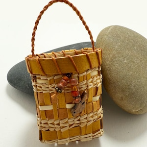 K&ouml;nnte beinhalten: Ein kleiner, handgefertigter Korb mit einem gewebten Design in Beige- und Hellbraunt&ouml;nen. Der Korb hat einen geflochtenen Griff und ist mit einem Perlenakzent und einem kleinen Holzvogel verziert. Der Korb ist etwa 7.6 cm hoch.