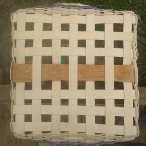 Op de afbeelding: Een geweven mand met een lichtbruine houten handvat. De mand is gemaakt van natuurlijke materialen en heeft een lichtbeige kleur. De mand is geweven in een rasterpatroon.