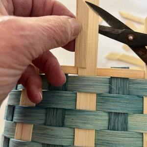 May include: A hand holds a piece of wood, cutting it with scissors to create a woven basket. The basket is made of natural coloured and blue stained bamboo.