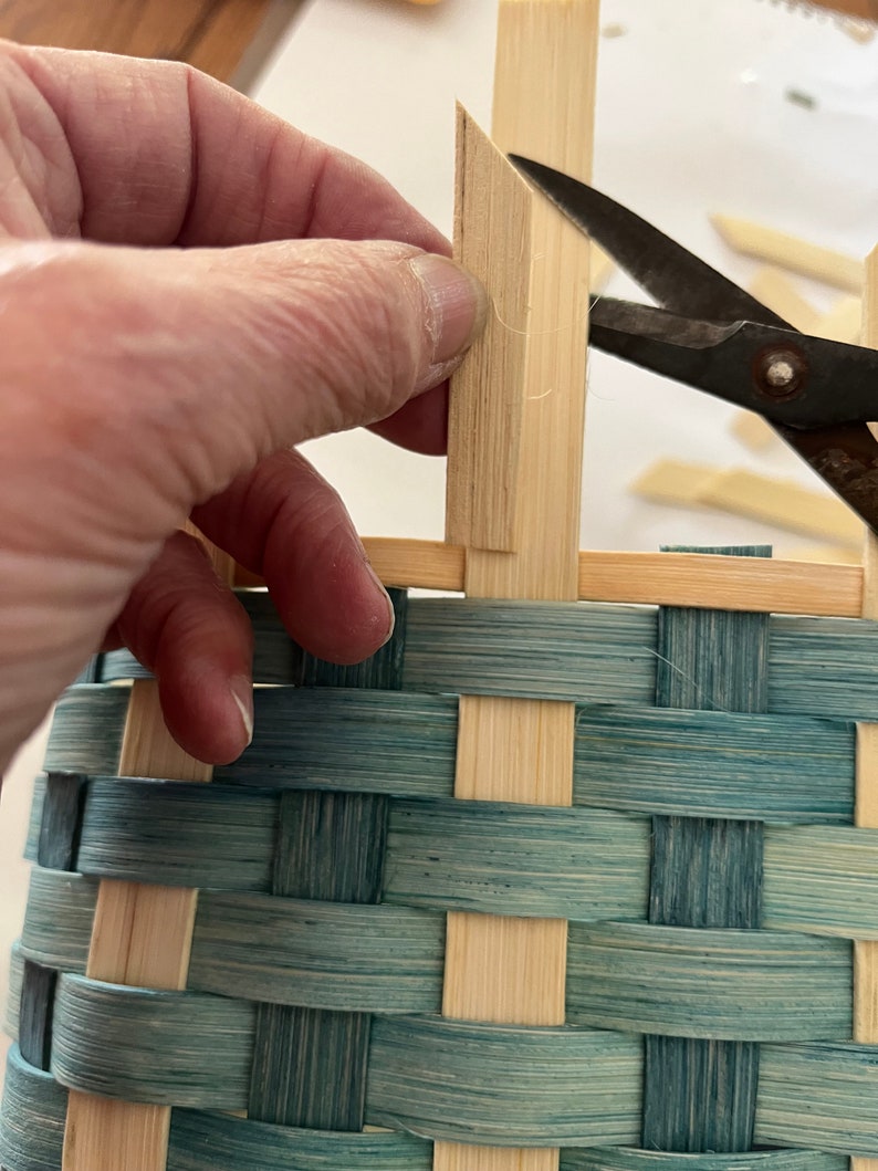 May include: A hand holds a piece of wood, cutting it with scissors to create a woven basket. The basket is made of natural coloured and blue stained bamboo.