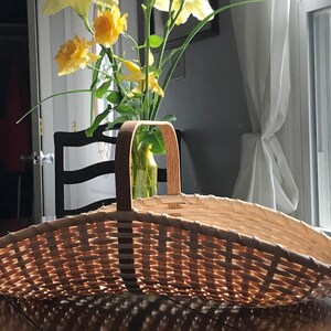 May include: A woven wicker basket with a wooden handle. The basket is empty and sitting on a wooden table.