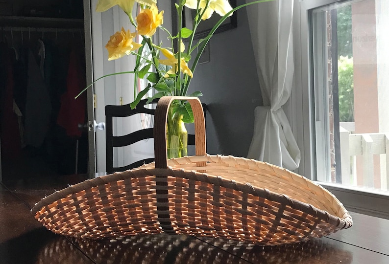 May include: A woven wicker basket with a wooden handle. The basket is empty and sitting on a wooden table.