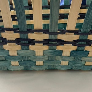 May include: Close-up of a woven basket with a light blue and natural coloured weave. The basket has a black rim and a black woven design around the top.