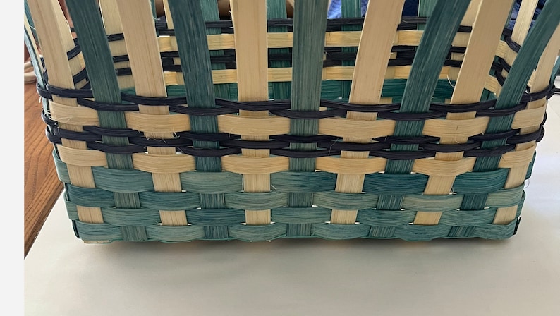 May include: Close-up of a woven basket with a light blue and natural coloured weave. The basket has a black rim and a black woven design around the top.