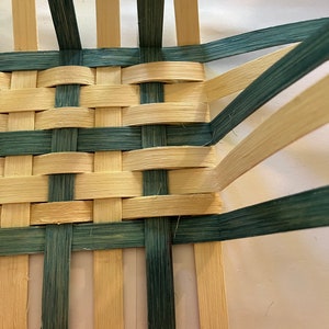 May include: Close-up of a woven basket with natural coloured and green coloured reeds. The basket is woven in a checkerboard pattern.
