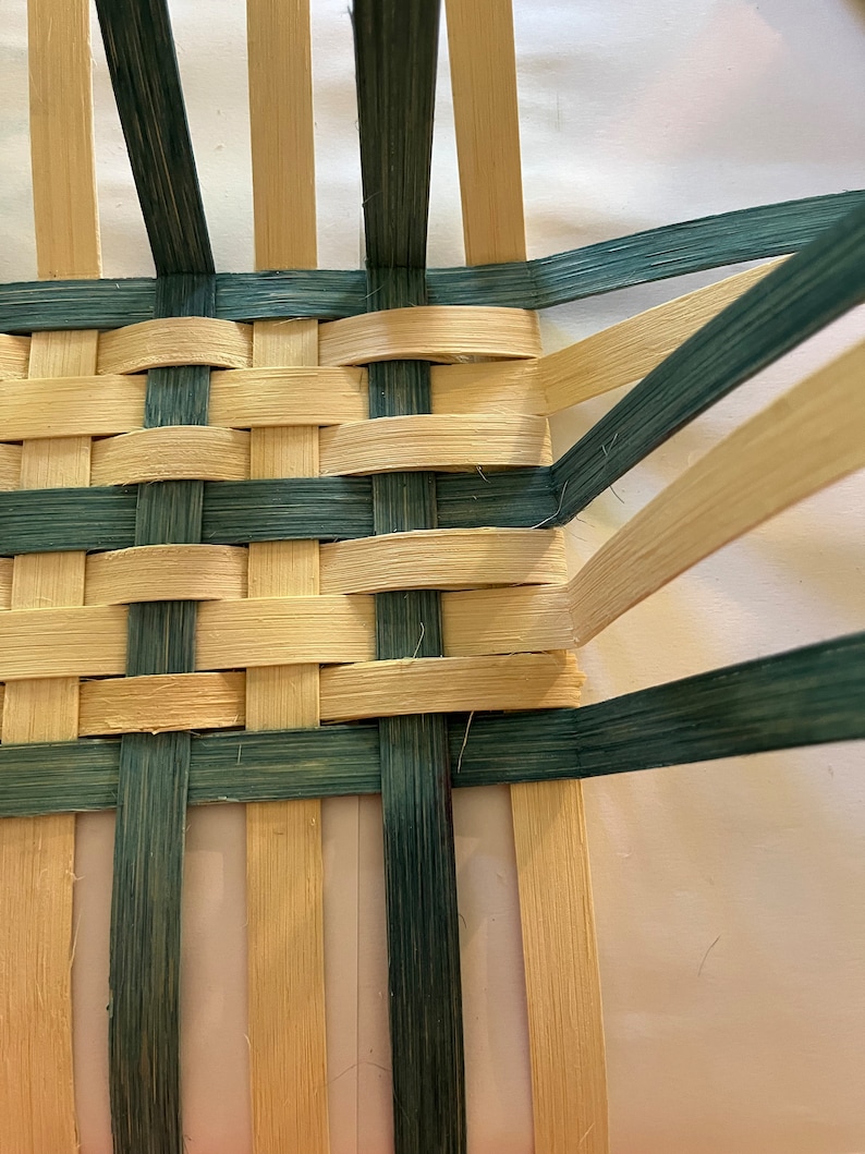 May include: Close-up of a woven basket with natural coloured and green coloured reeds. The basket is woven in a checkerboard pattern.