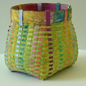 May include: A woven basket with a green, pink, yellow, and blue pattern. The basket is made of paper and has a square base.