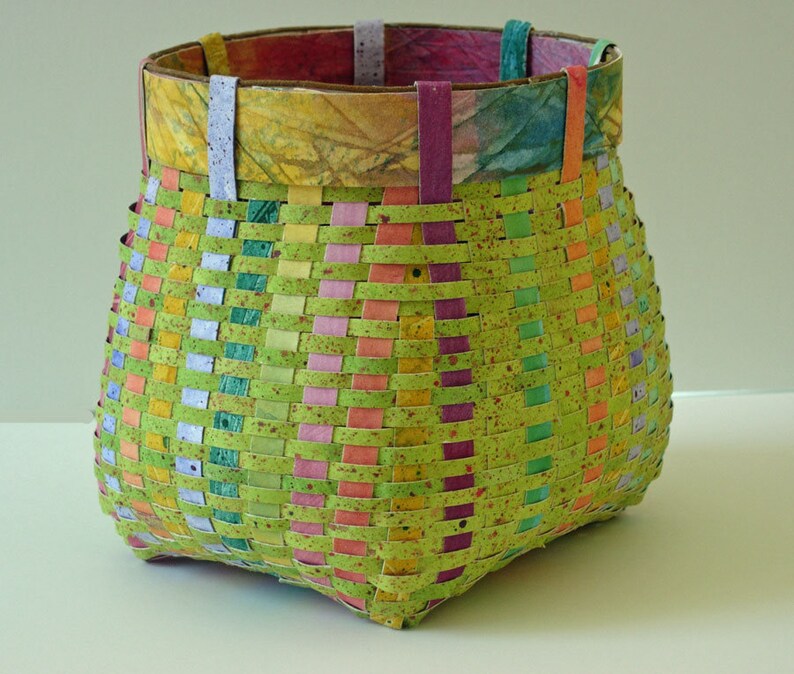 May include: A woven basket with a green, pink, yellow, and blue pattern. The basket is made of paper and has a square base.