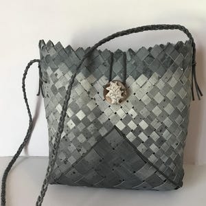 Op de afbeelding: Een grijze geweven handtas met een gevlochten riem en een decoratieve hanger. De tas heeft een geometrisch patroon en een geschulpte bovenrand. De hanger is lichtbruin met een wit bloemmotief.