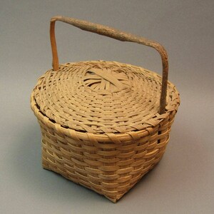 May include: A small, round, woven basket with a lid and a wooden handle. The basket is made of natural materials and has a light brown colour.